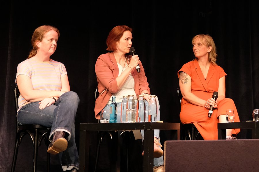 Drei Frauen sitzen nebeneinander auf einer Bühne. Eine spricht in ein Mikrofon.