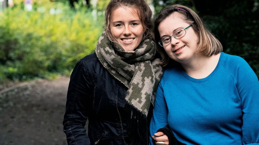 Zwei junge Frauen mit braunen Haaren in die Kamera lächelnd. Die Rechte trägt ein blaues Oberteil und eine Brille, die Linke ein schwarzes Oberteil und einen grün und grau gemusterten Schal. 