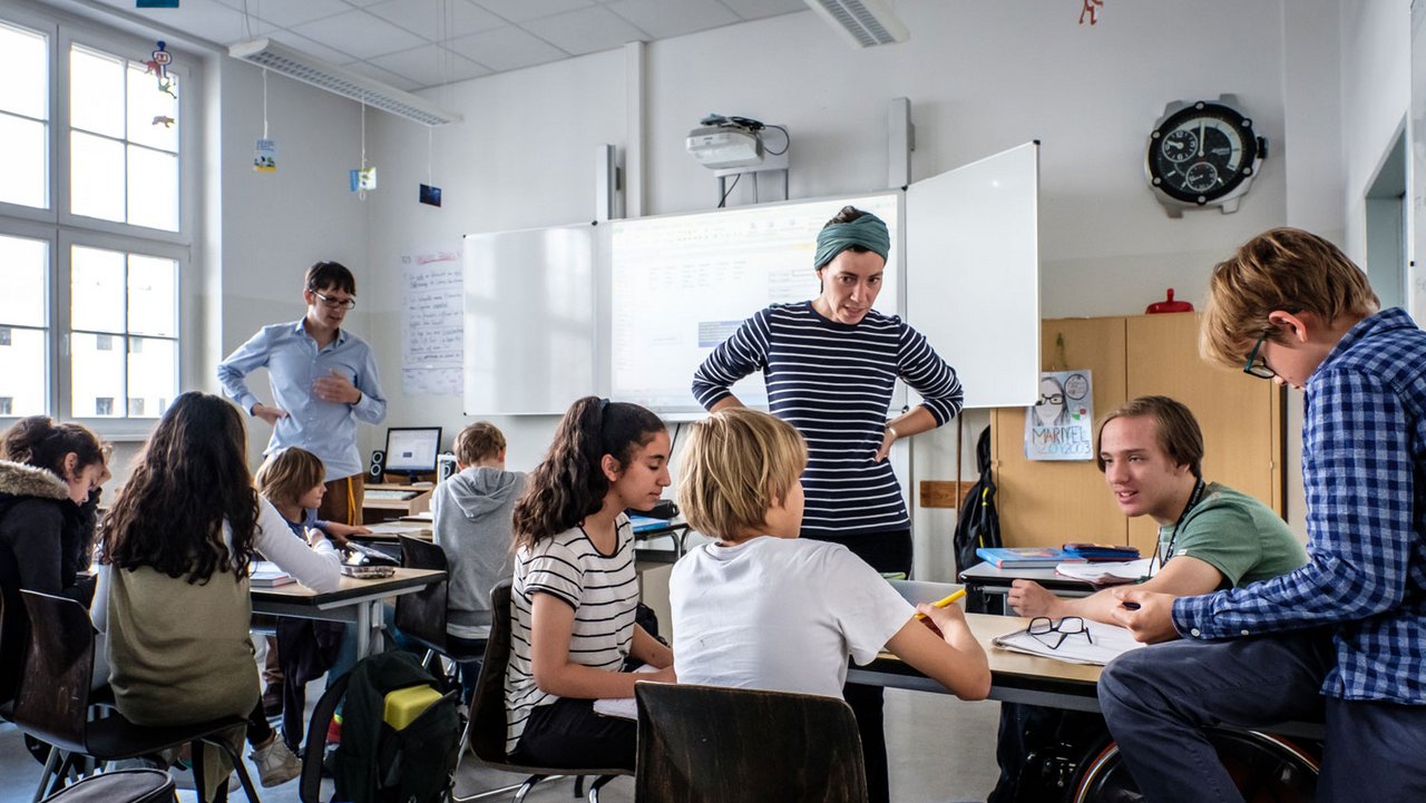 Etwa 10 Schüler in einem Klassenzimmer an Tischen sitzend. Zwei Lehrer erklären ihnen etwas. Im Hintergrund ein großes Whiteboard. 