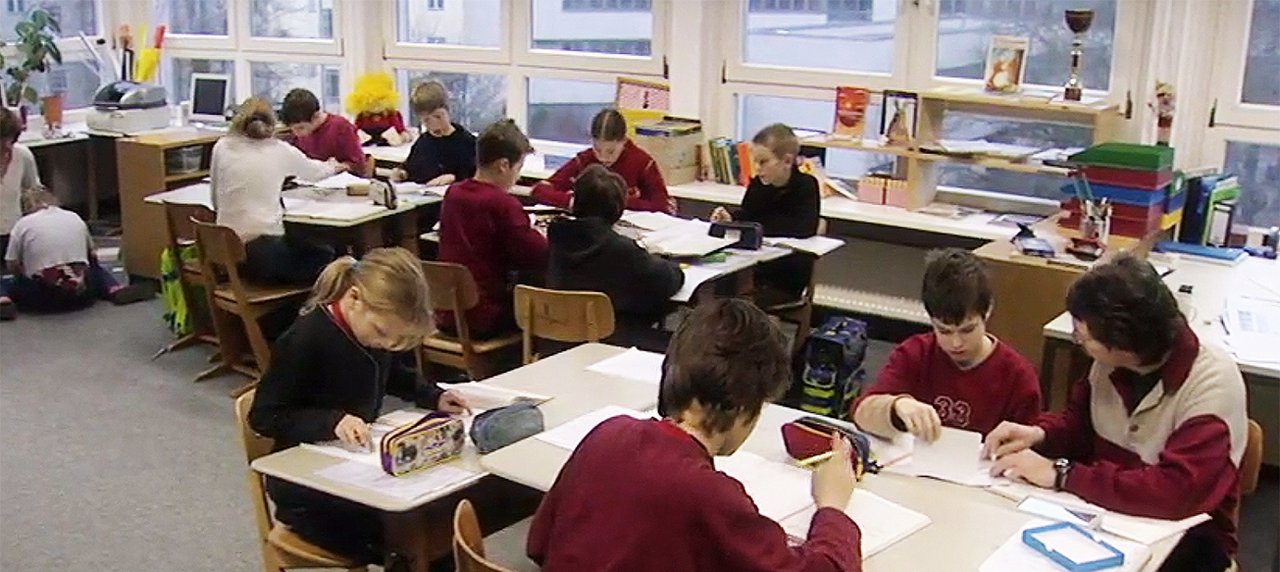 Etwa 10 Kinder im einem Klassenzimmer an Gruppentischen sitzend. Sie sind jeweils über ein Blatt Papier gebeugt.