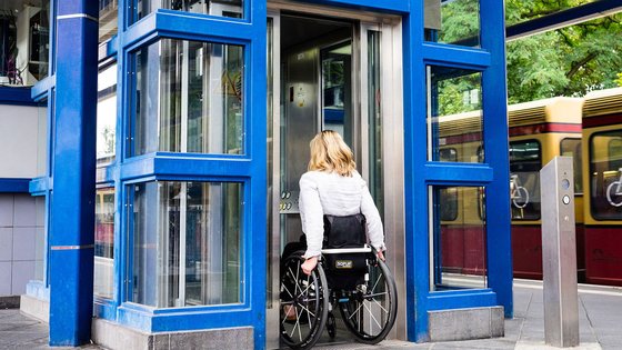 Eine blonde Frau im Rollstuhl steht vor einem Aufzug. Sie trägt ein weißes Oberteil.