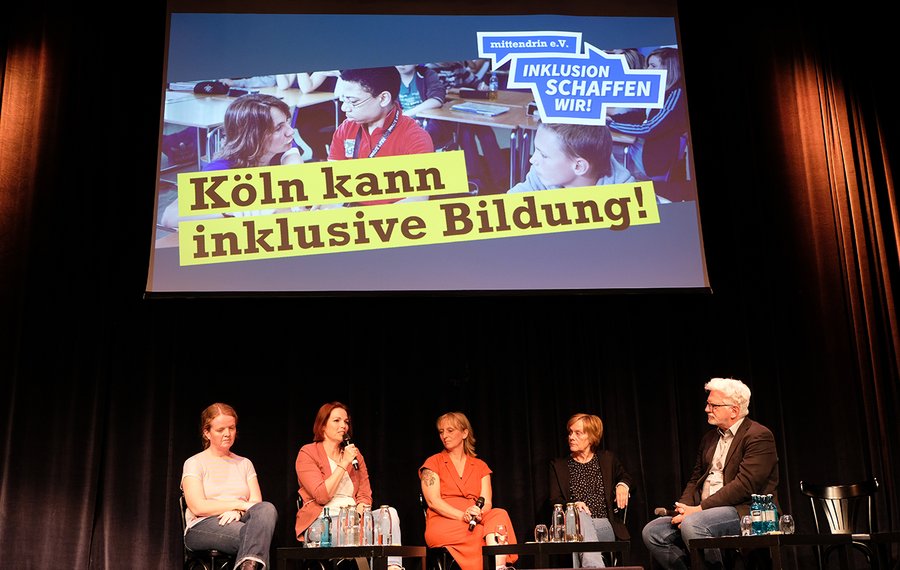 Vier Personen sitzen nebeneinander auf einer Bühne. Über Ihnen ist ein Bild in den Hintergund projiziert. Auf gelben Balken steht in schwarzer Schrift „Köln kann inklusive Bildung!" Darüber steht das Logo des mittendrin e.V. mit der Unterzeile „Inklusion schaffen wir!"