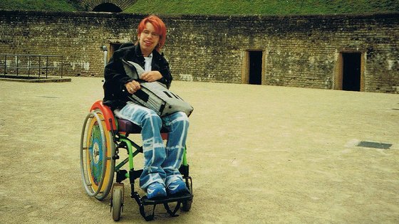 Eine junge Person mit roten Haaren im Rollstuhl. Sie trägt ein dunkles Oberteil und eine Jeanshose und hält eine Tasche auf dem Schoß.