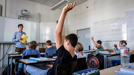 Schüler in einem Klassenzimmer. Ein Schüler mit schwarzem Oberteil und schwarzen Haaren hat den Arm gehoben.