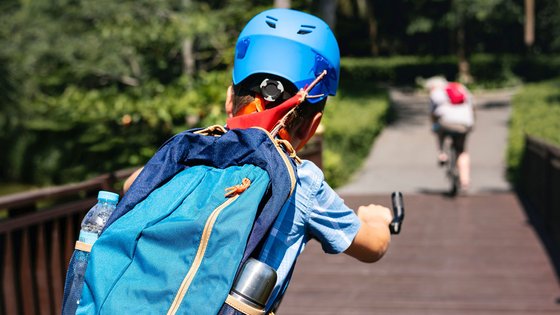 Ein Kind mit blauem Helm und blauem Rucksack auf einem Fahrrad fahrend. Es hat dem Betrachter den Rücken zugewandt. 